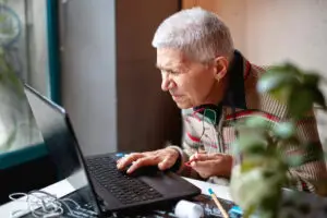 Eine ältere Frau sitzt vor einem Laptop und kann ganz offensichtlich die Inhalte schlecht erkennen. Barrierefreiheit für Websites kann helfen.